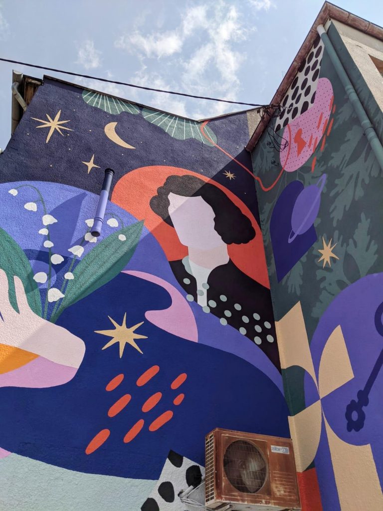 Kolorowy mural na ścianie budynku z postacią i roślinnymi motywami.