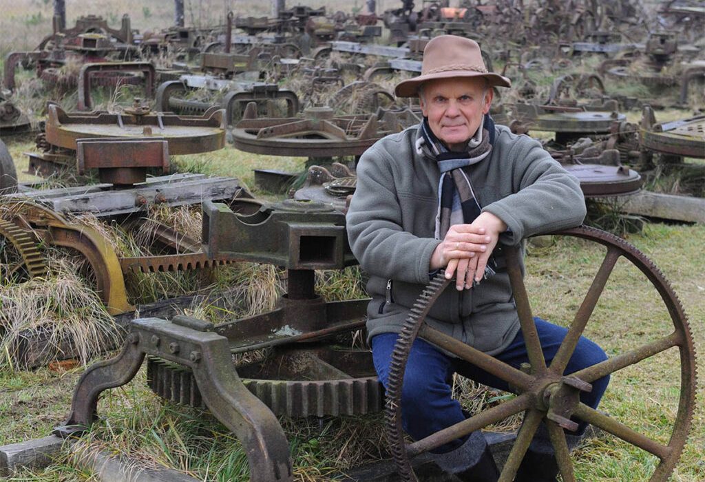 Mężczyzna siedzący przy starym kole zębatego wśród zardzewiałych maszyn rolniczych.