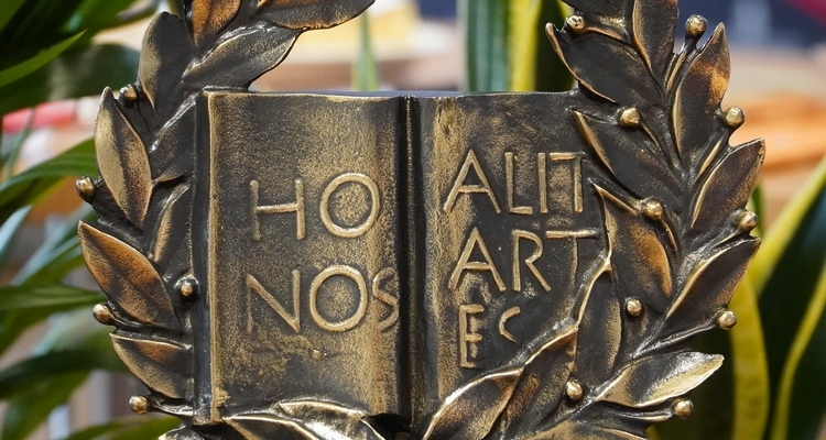 Złota statuetka w kształcie otwartej książki otoczona liśćmi.